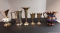 Lot of six brass and metal cups, vases, and pitchers in a row against a plain background.