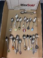 Top-down image of approximately 25 collector spoons arranged in a cardboard box with a MaxSold measurement guide.
