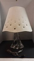Crystal table lamp with white fabric lampshade decorated with circular cutouts, showing lamp as lit from front perspective.