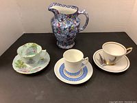 Photo showing three cups and saucers and a blue floral jug arranged on a black surface with a white background.