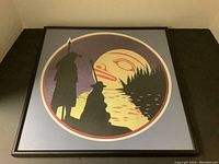 Framed artwork showing two figures silhouetted against a moonlit water landscape with trees and a stylized moon face.