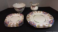Photo showing 4 small square plates stacked, 2 large round plates under, small bowl and pitcher in back