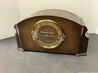 Front view of the wooden mantel clock showing its shape and wood finish with brass bezel and glass cover.