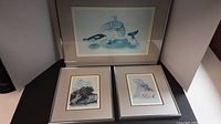 Three prints by Sue Coleman shown: two smaller 5x7" prints of Seal and Bear with frames and matting, laid flat. Larger 15x10" Orca print framed, leaning on wall.