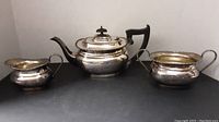Front view of the silver plated teapot, creamer and sugar bowl on a dark surface.