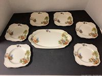 Seven-piece dish set on black background. Includes a large rectangular serving plate in the center surrounded by six smaller side plates. Each piece has floral and cottage scene decoration with scalloped edges.