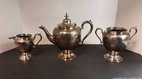 Front view of three silver plated items: tea pot, pitcher, and double handled vase with ornate designs.