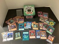Photo showing the Yugi-oh trading cards arranged around the branded tin box. Cards include a mix of monster, spell, and trap cards with various colors and protective sleeves.