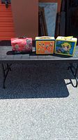 Three vintage metal lunch pails displayed on outdoor table: Pro Sports, Zelda Nintendo DS, and Dale Earnhardt themed.