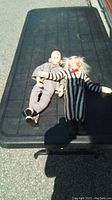 Two dolls laid flat on a black surface outdoors, showing full bodies of Pee Wee Herman and Beetlejuice dolls.