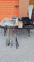 Wide view of lot showing six fishing rods leaning against table and multiple tackle boxes on table.