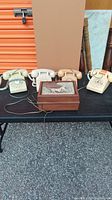 Five vintage telephones displayed on a black table with a wooden decorative box in front, showing rotary dial designs and coiled cords.