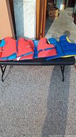 Three adult life jackets displayed on a bench outdoors, two red with blue accents, one blue with yellow detailing.