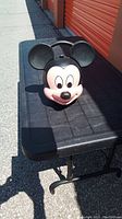 Front view of lunch pail shaped like Mickey Mouse head on black table outside.