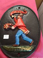 Close-up of oval cast iron fireman sign showing side profile of fireman in red coat blowing trumpet with lit torch in hand, mounted on black background with red banner labeled 'FIRE'.