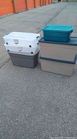 Six plastic storage totes stacked outside on pavement, including white, beige, and turquoise colors.