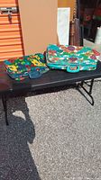 Two brightly colored floral pattern vintage suitcases on a folding table outside in sunlight, background includes storage unit door and wall