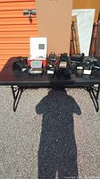 Table display of multiple vintage cameras and accessories including flash units and cases.