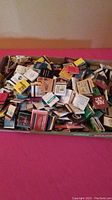 Box filled with a large pile of assorted vintage matchbooks in varied colors and designs.
