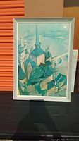 Full view of the framed cubist style art print showing the abstract cityscape with buildings and church steeple in teal and beige tones.