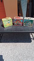 Photo of three vintage Disney metal lunch boxes: Snow White, Mickey Mouse Club, and Disney Express, displayed on a black table outdoors.