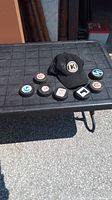 Seven vintage hockey pucks arranged on a black table surface outdoors along with a black baseball hat featuring Kingston Frontenacs logo.