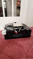 Black Sony AV receiver with cables and manual on top, placed on carpeted floor in room with white walls and window.