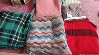 Six blankets and throws displayed on pink carpet including Hudsons Bay wool blanket, plaid blanket, patterned blanket in plastic, pink throw, beige textile and table covering.