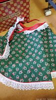 Green Christmas apron with candy cane print and white lace edges on floor next to box with Hickory Farms label
