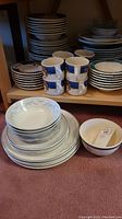 Wide view of stacked plates, bowls, and mugs on shelf and floor showing various patterns and sizes
