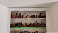 Wide photo showing 13 wicker duck-shaped baskets on white shelves: various sizes and styles of woven duck baskets are displayed, some with handles and some plain.