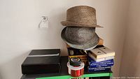 Photo showing four hats stacked on top of boxes, including hats in gray and beige tones along with boxes of playing cards and poker chips
