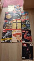 Photo of 10 vintage magazines including 9 Life mags, 1 Time mag, arranged on floor with various covers visible showing different topics and imagery.