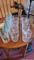 Eight etched clear glass cups in metal holders arranged on a metal wire carry tray with coil handle, shown with five additional blue and white striped glasses on a wooden table.