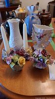 Overview of porcelain flowers, candle holders with white tapers, and three vases on wooden surface