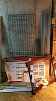 Two unassembled wire grid metal shelves and a partially visible Husky box with storage unit packaging against a concrete wall.