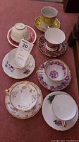 Seven porcelain teacups and saucers arranged on carpet, shows varying floral and gold accent decoration.