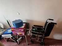 Photo showing the Airgo wheelchair, two shower chairs, box of Kirkland adult diapers, small blue padded item on top, part of bed railing visible