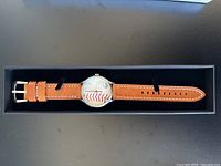 Watch face showing baseball seam dial and brown leather strap inside presentation box
