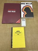 Overview of three items: red hardcover, white biography, yellow tour booklet