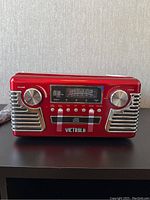Front view of red Victrola AM/FM radio with volume and tuning knobs, digital display, and buttons below.
