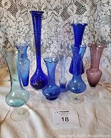 Eight assorted glass bud vases mostly in shades of blue with one amethyst piece, arranged on a lace background. Various shapes and frilly rims are visible.
