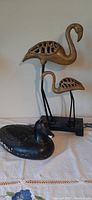 Photo showing two carved flamingo figurines on metal legs and a black wooden duck decoy on cloth surface.