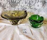 Two handcrafted green glass bowls displayed side by side on a white cloth with a lace curtain in the background. One is dark green and abstract, the other bright green and rounded.