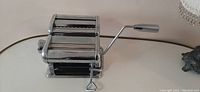 Pasta maker machine with hand crank attached, showing rollers and cutter mechanisms.