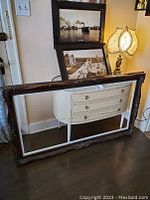 Large empty ornately carved dark wood picture frame with white interior, leaning against a wall and floor, showing detailed wood work and beveled edge.