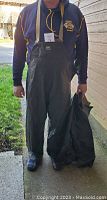 Photo showing a person wearing the black bib-style rain pants with adjustable suspenders, held up by the person standing outside, showing the size fit and front Helly Hansen logo patch.