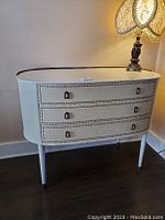 Full front view of small console table showing the triple drawers, decorative nail accents around drawer edges, brass rail on top surface, and tapered legs with brass feet.
