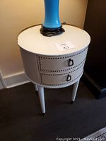 Full side view of the small round side table with double drawers, white top with brass rail, decorative nailhead detail, and tapered legs.
