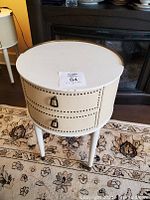 Full front view of the small round white end table showing two drawers with ring pull handles and decorative nail detail on vinyl surface.